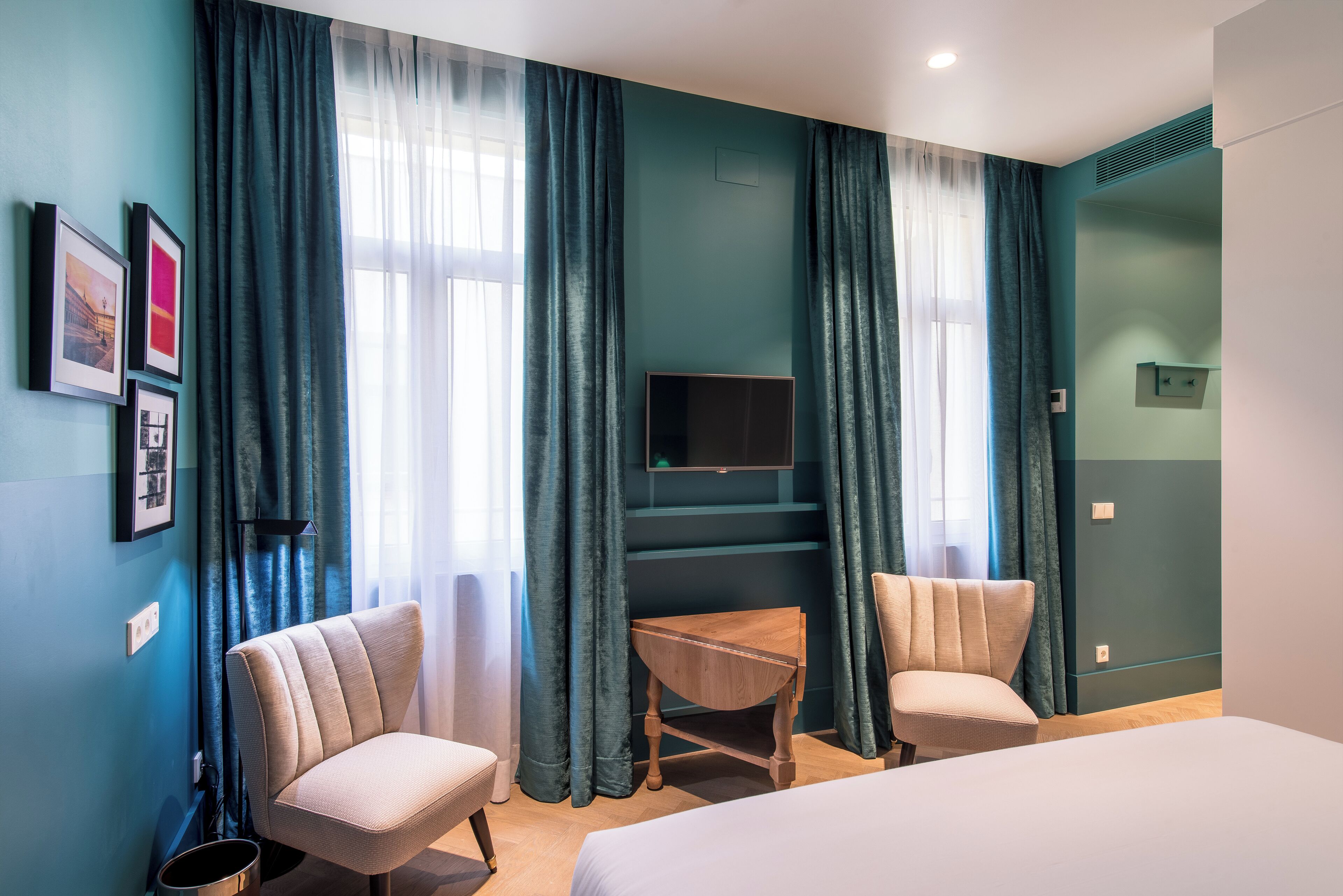 A hotel room with teal walls, a bed, two armchairs, a wooden side table, a flat-screen TV, and a window with sheer curtains.