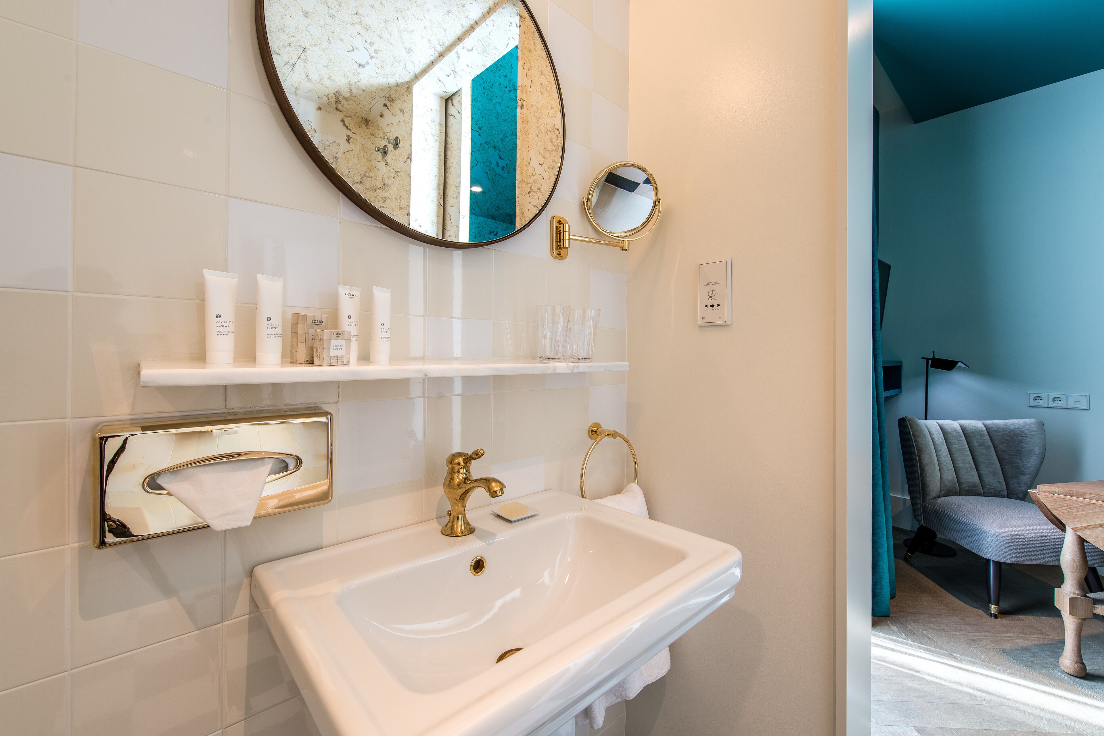 Un baño con lavamanos blanco, detalles dorados, un espejo redondo y estante con artículos de tocador.