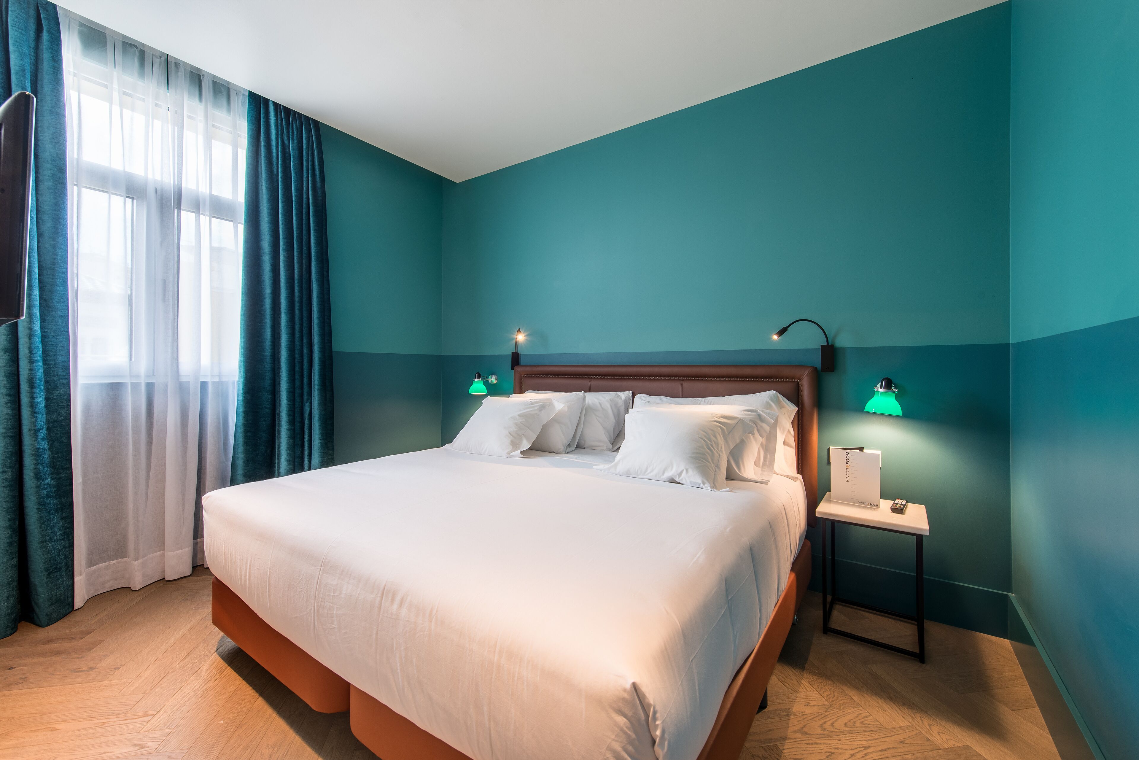 Una cama bien hecha en una habitación con paredes color turquesa, suelo de madera y dos lámparas de noche.