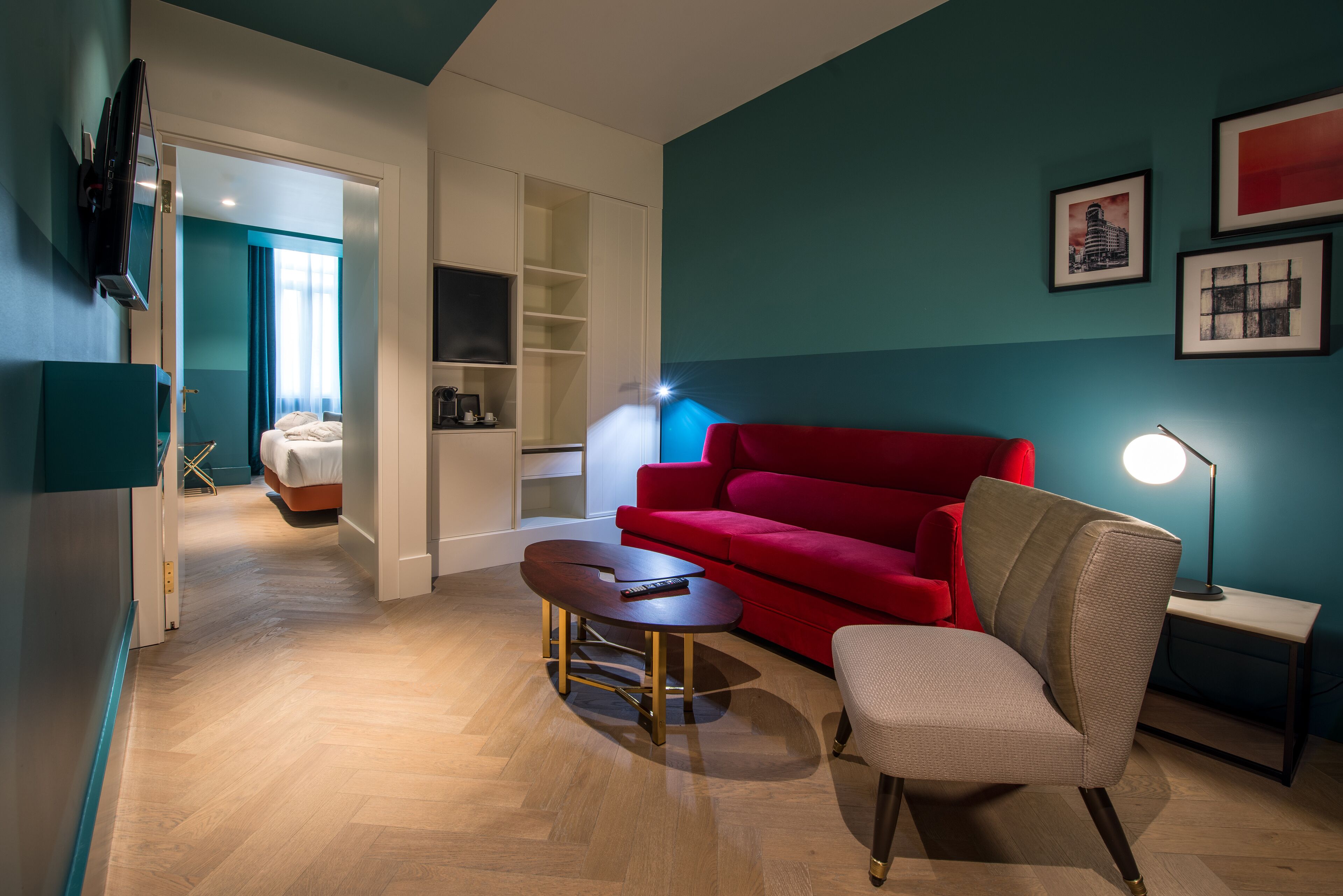 A modern hotel room with a red sofa, a small round table, a lamp, and a TV mounted on the wall.