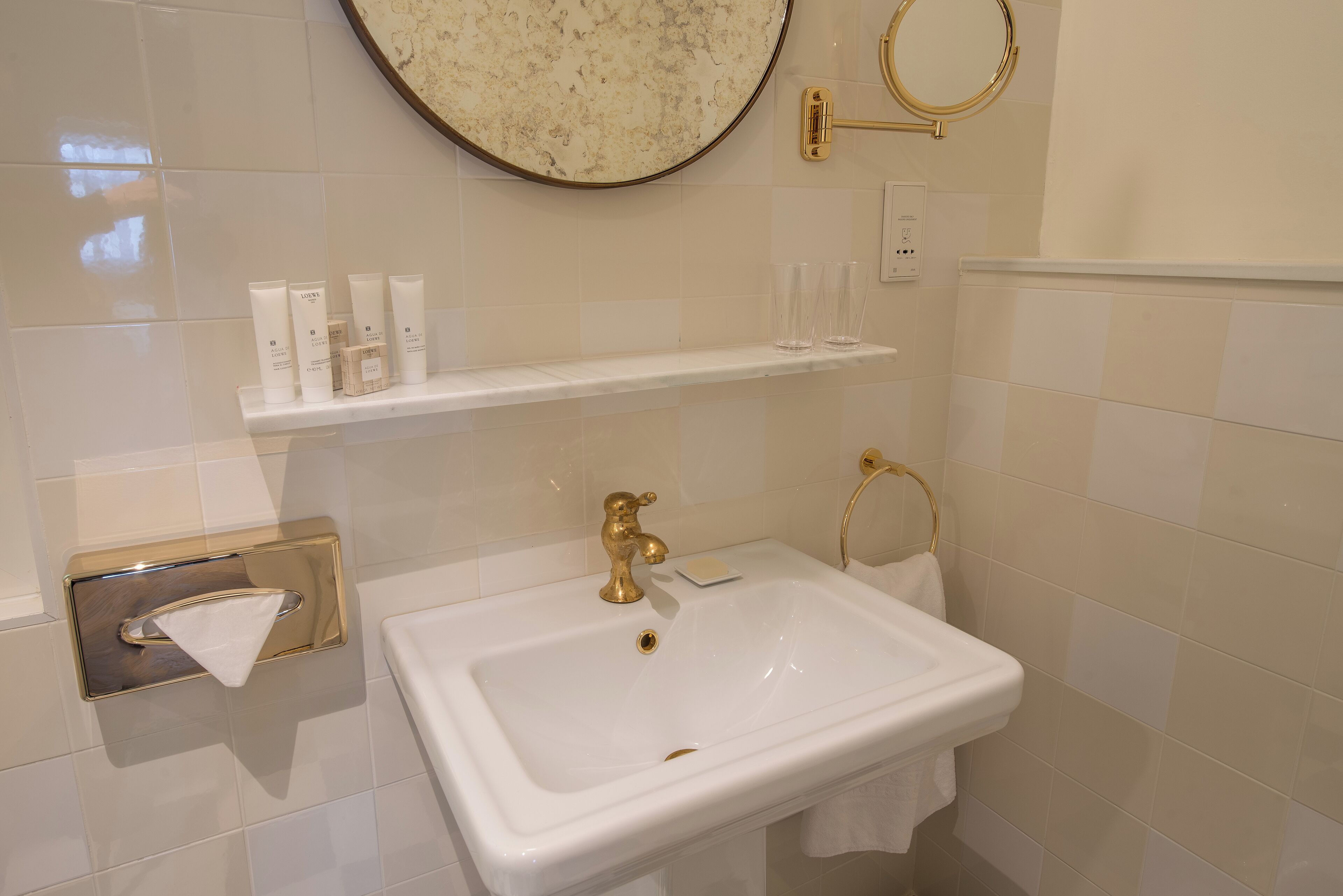 Casa de banho com lavatório branco, acessórios dourados, um porta-toalhas e uma prateleira com artigos de higiene pessoal.