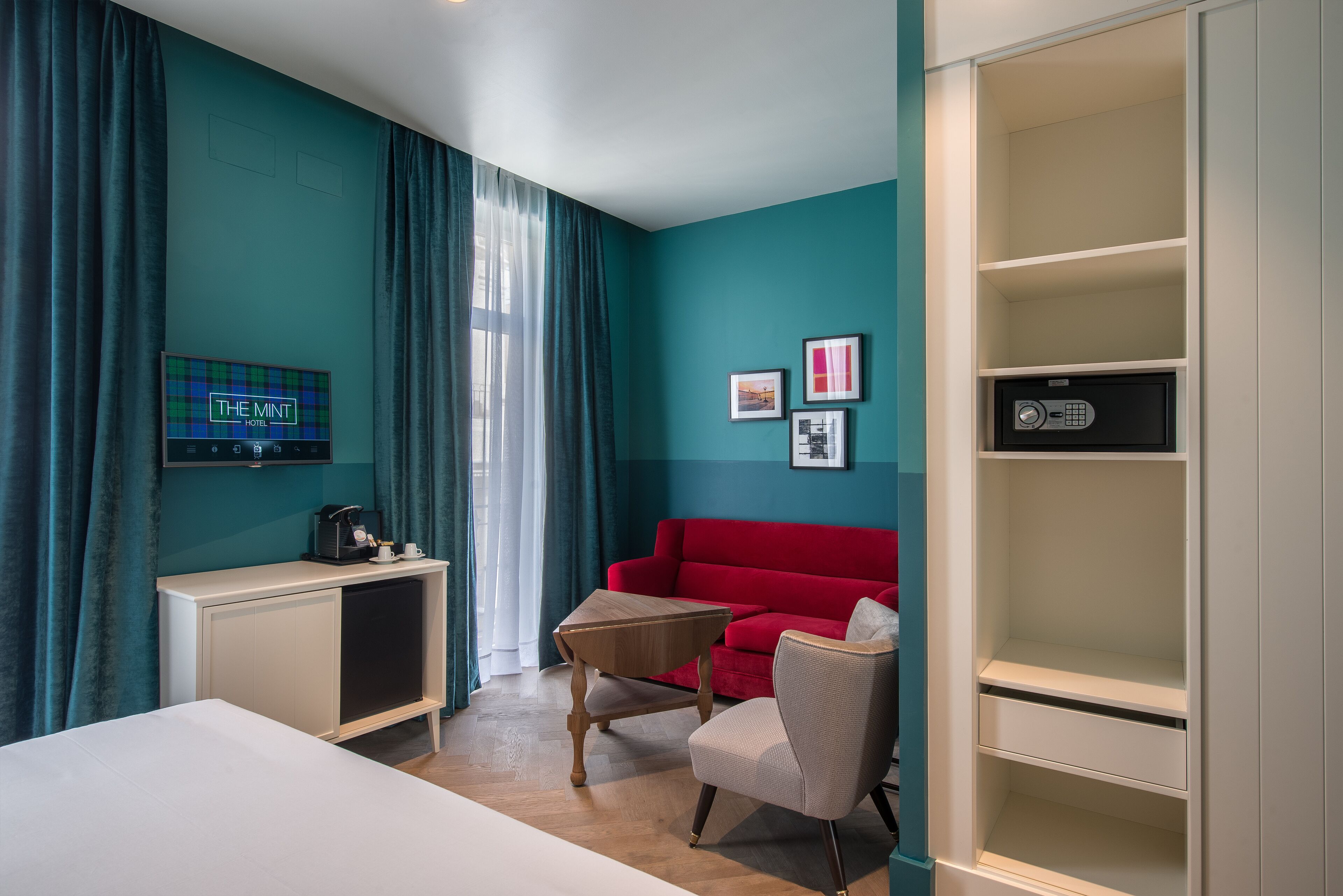 Habitación de hotel con paredes verde azulado, un sofá rojo, una mesita de madera, un armario blanco y una nevera minibar.
