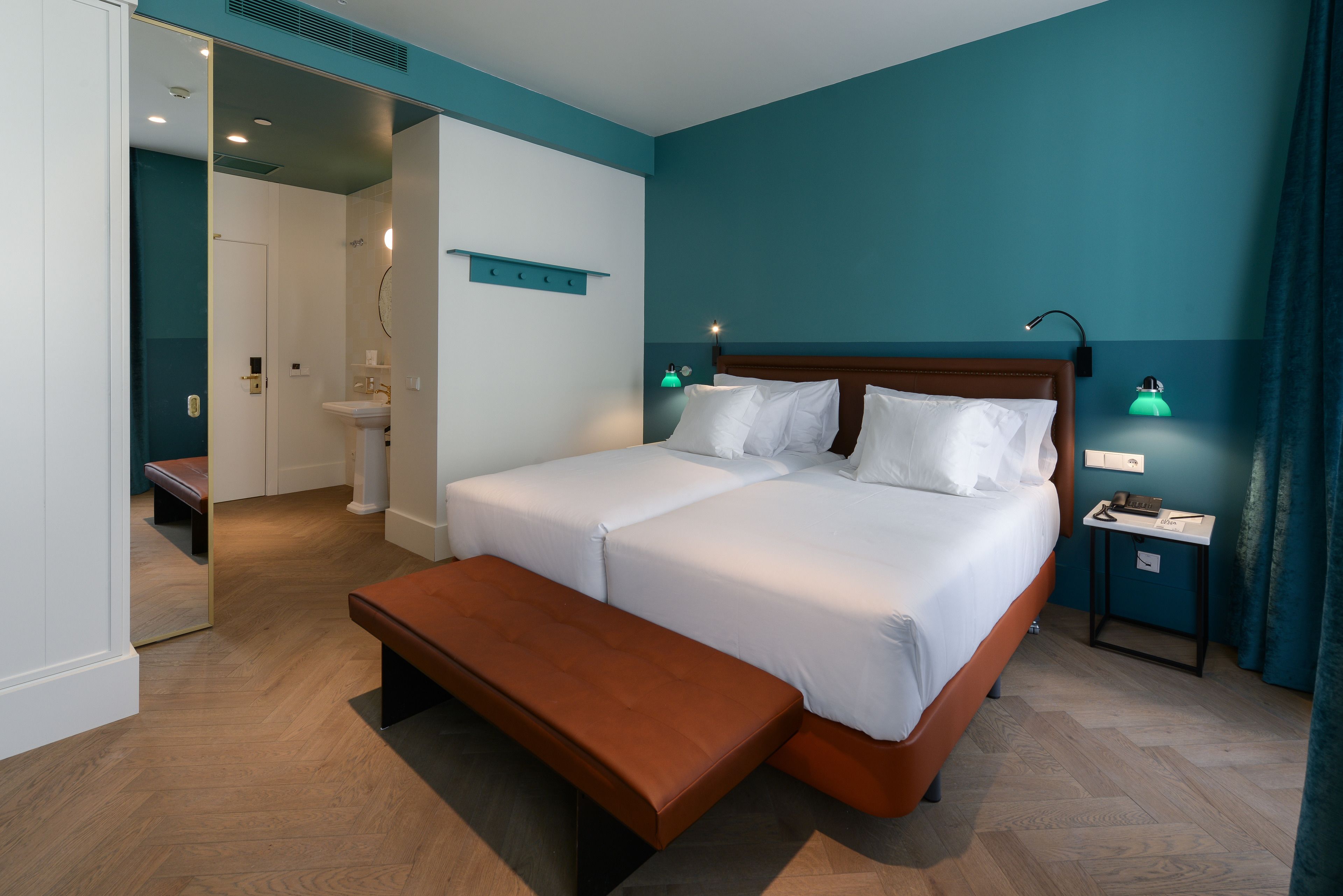 Una cama doble con sábanas blancas, un banco de madera, paredes color turquesa y una mesita de noche con una lámpara.