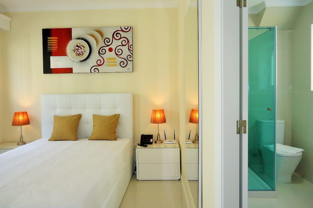 A hotel room with a bed, bedside lamps, a nightstand, a wall art piece, and a bathroom visible through an open door.