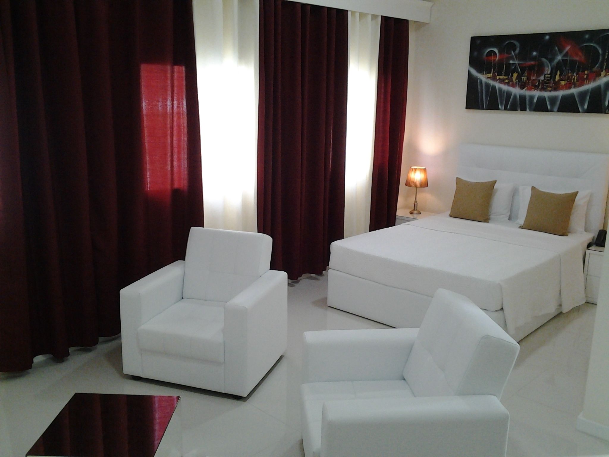 A hotel room with a bed, two white armchairs, a nightstand with a lamp, and a painting on the wall.