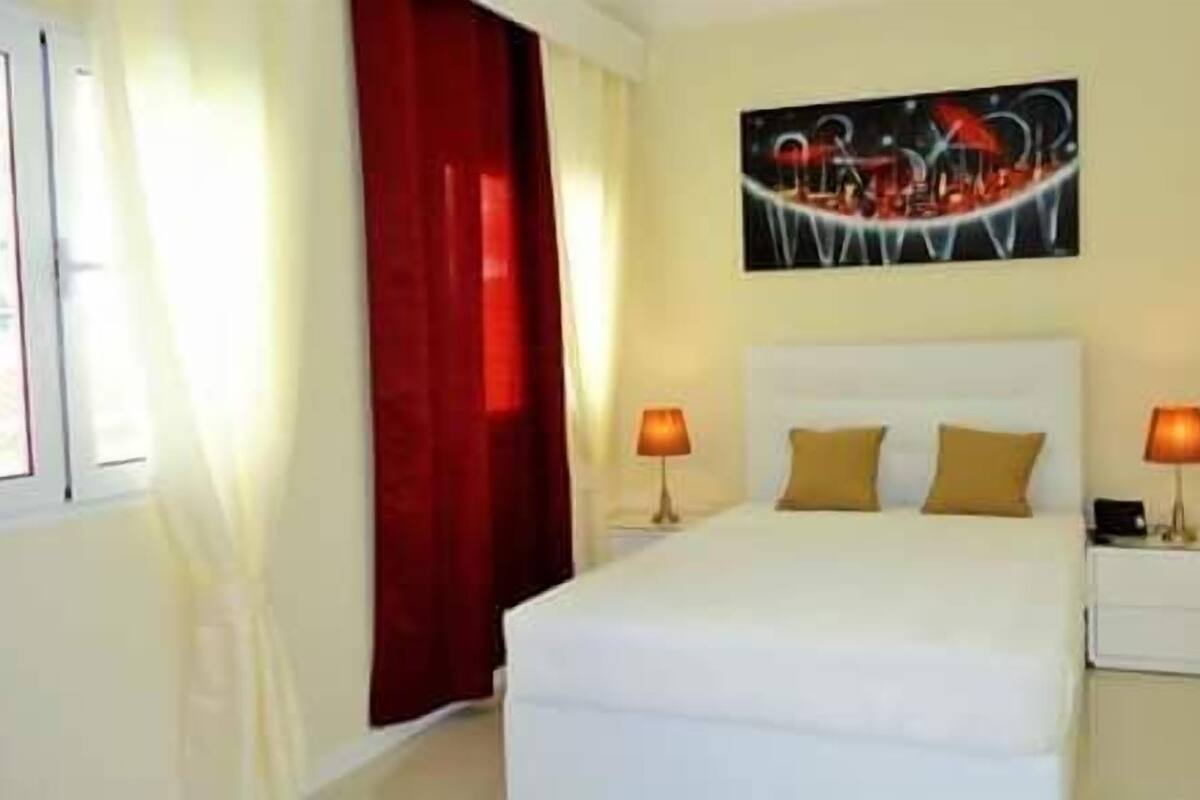 A hotel room with a bed, two bedside tables with lamps, a red curtain, and a painting on the wall.