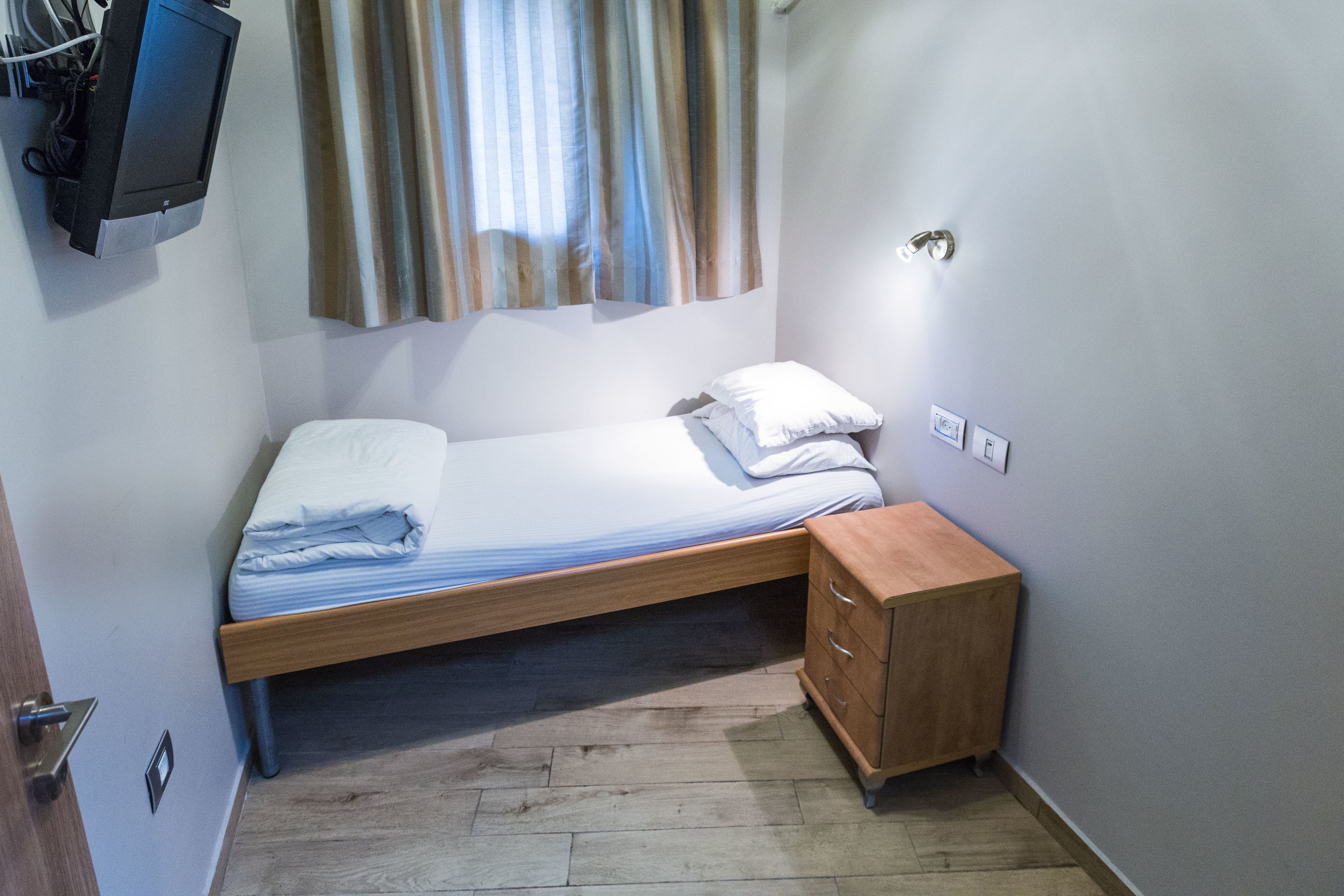 Una cama pequeña y bien hecha con estructura de madera, una mesita de noche con cajones y un televisor montado en la pared.