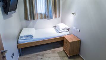 Una cama pequeña y bien hecha con estructura de madera, una mesita de noche con cajones y un televisor montado en la pared.