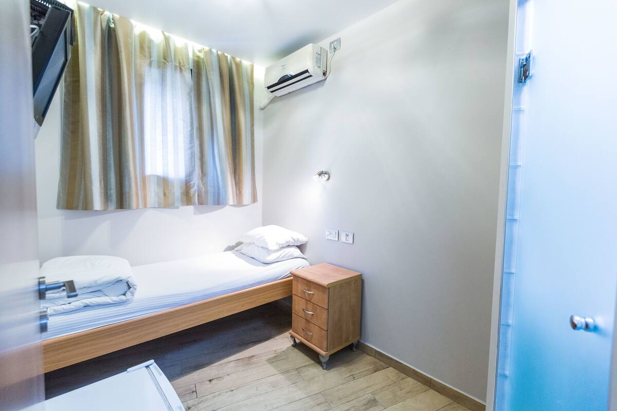 Una habitación pequeña y limpia con una cama individual, un aparador de madera y un equipo de aire acondicionado instalado en la pared.