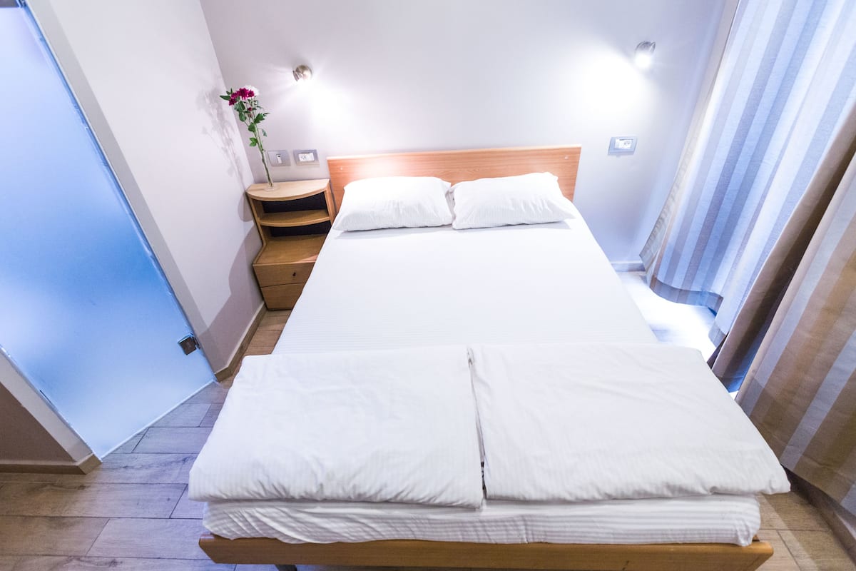 Una cama con ropa blanca impecable, cabecera de madera y mesita de noche con un jarrón de flores.