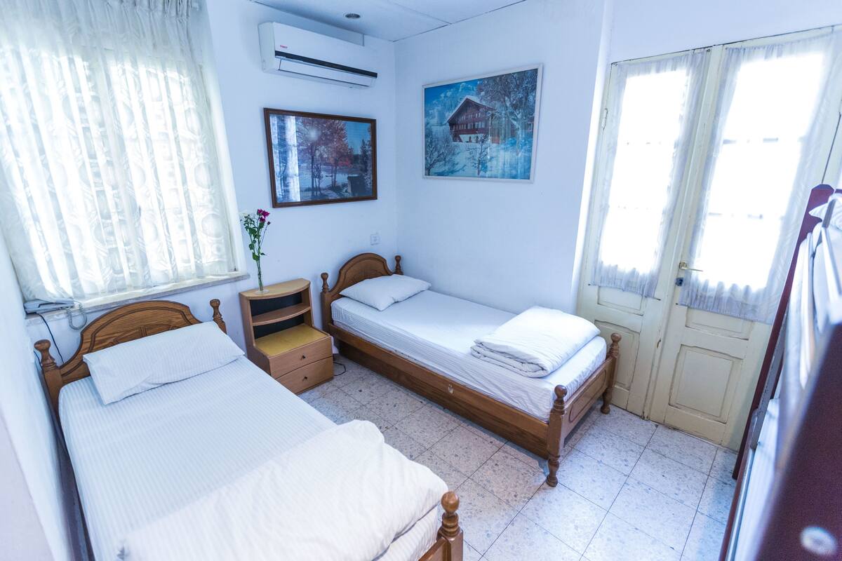 Un dormitorio con dos camas individuales, una mesita de noche, una ventana con cortinas traslúcidas y un aire acondicionado de pared.