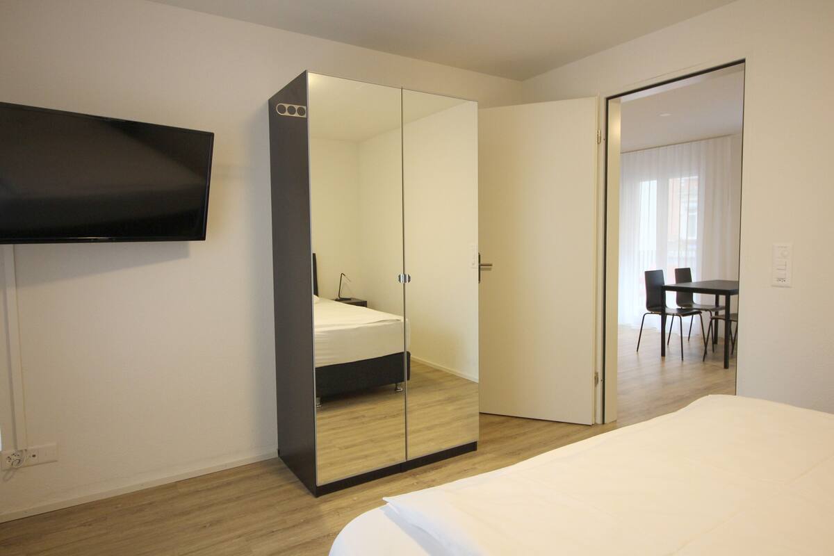 A hotel room with a bed, a wardrobe with a mirror, a television, and a dining area with chairs and a table.