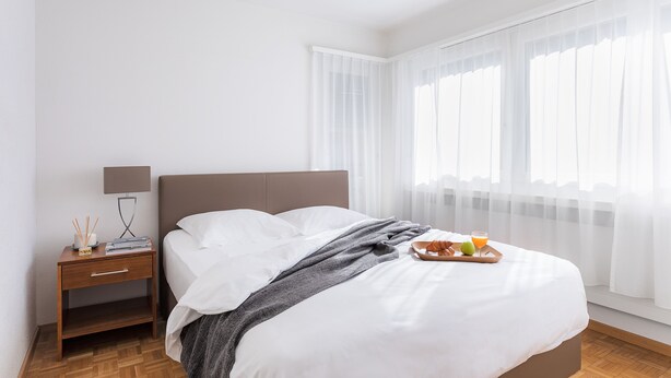 Uma cama bem arrumada com lençóis brancos, uma mesa de cabeceira com abajur e um tabuleiro com croissants e suco.