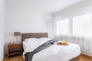 A neatly made bed with white linens, a bedside table with a lamp, and a tray with croissants and juice. - Swiss Star Wiedikon (Zürich)