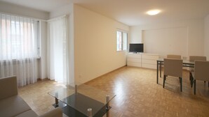 Ampla sala de estar com mesa de centro de vidro, sofá, mesa de jantar com cadeiras e televisão no canto.