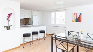 Uma cozinha moderna com armários brancos, forno embutido e janela com vista para a cidade. Três banquetas pretas e uma mesa de jantar de vidro com cadeiras.