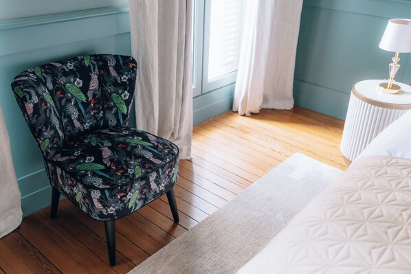 Una habitación con un sillón estampado con motivos florales, una cama blanca, una mesita redonda con una lámpara y una ventana con cortinas.