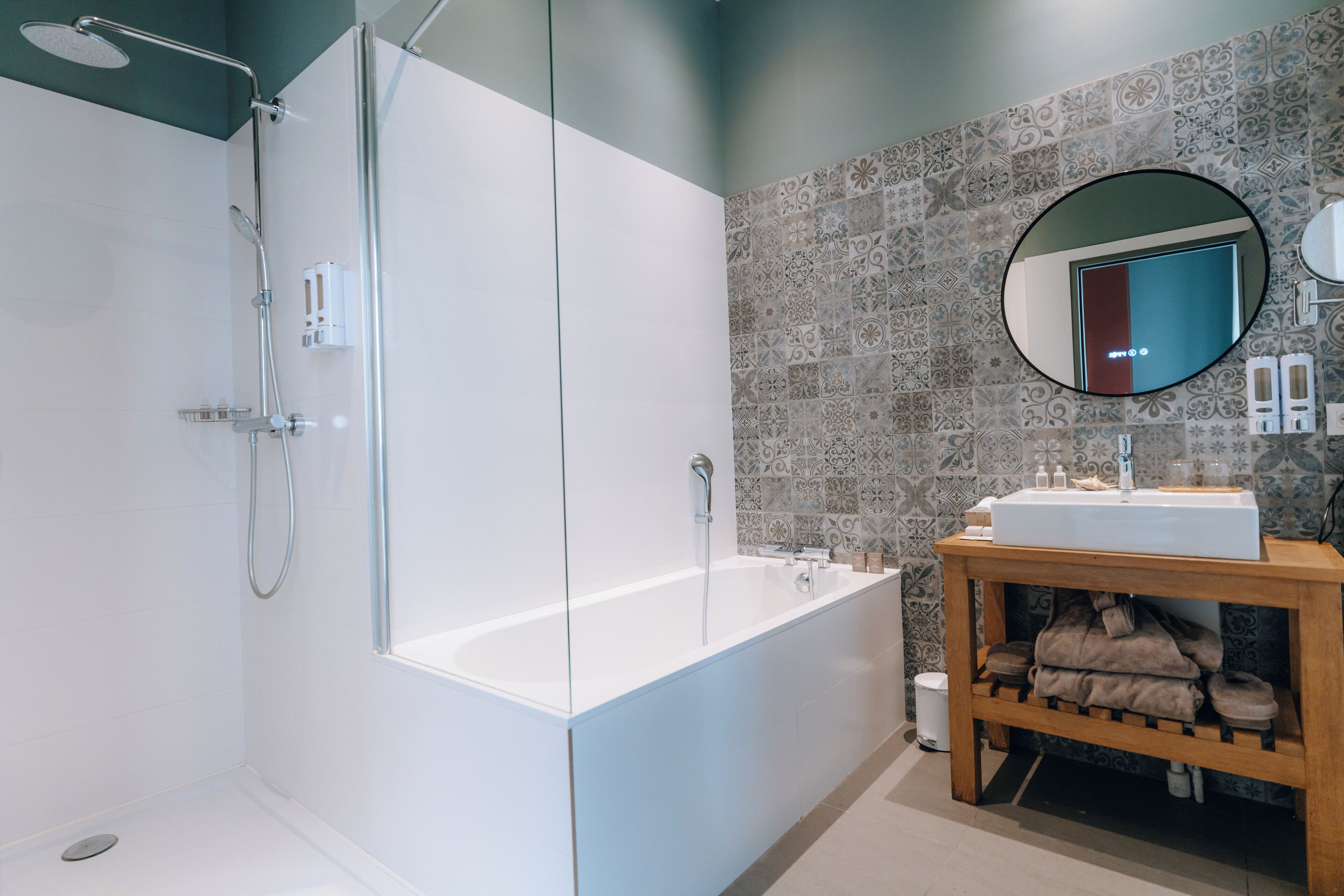 A modern bathroom with a glass-enclosed shower, a white bathtub, a wooden vanity with a sink, and patterned tiles on the walls.