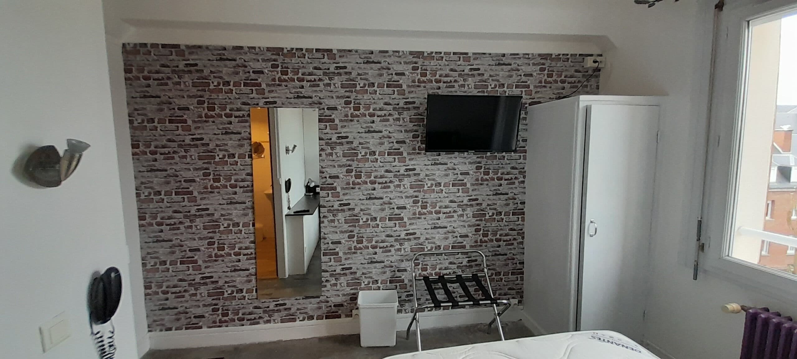 A room with a brick-patterned wall, a mirror, a television mounted above it, a wardrobe, and a radiator.
