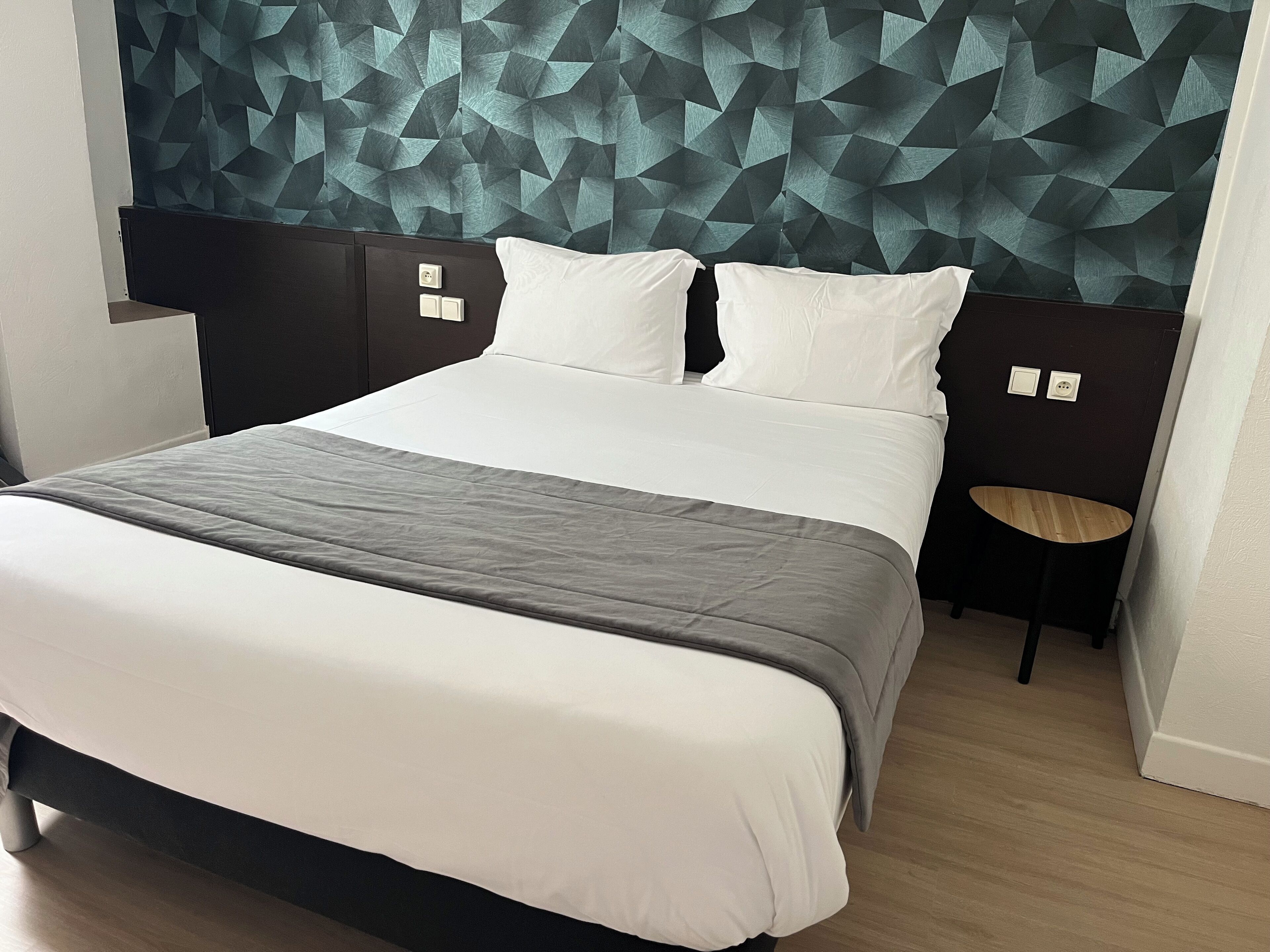 Una habitación de hotel moderna con un cabecero de estampado geométrico, una cama bien hecha con ropa de cama blanca y gris, y una pequeña mesita de madera.