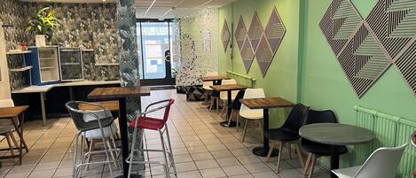 A cafe interior with a green wall, patterned wallpaper, and geometric wall art. Tables and chairs are arranged for seating.