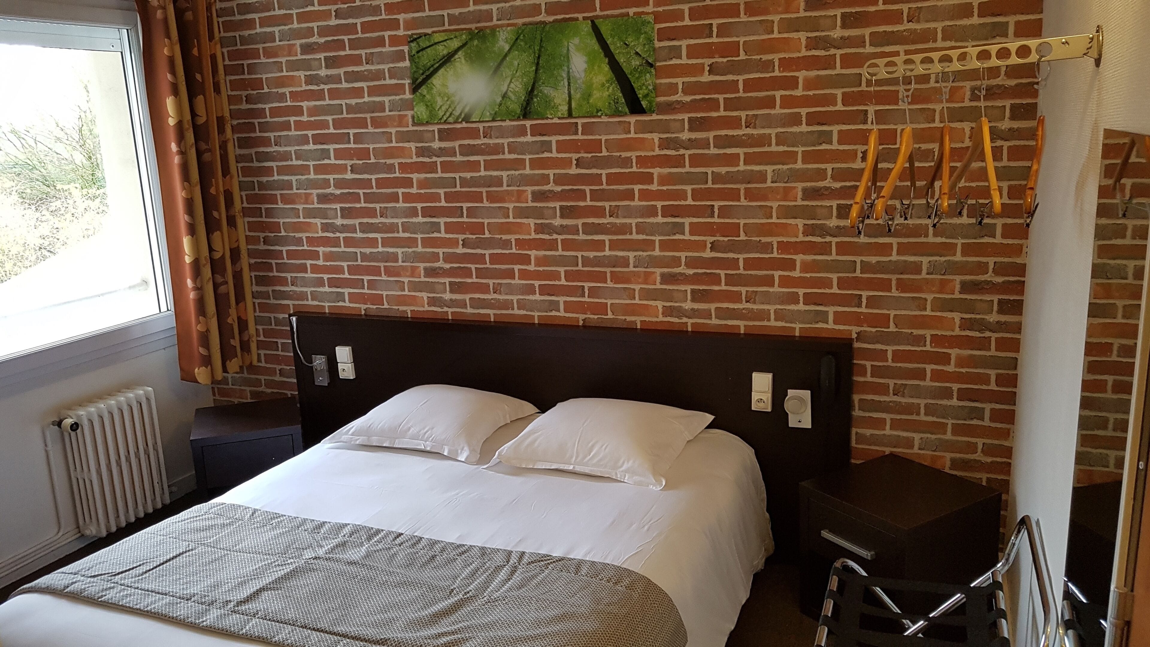 A hotel room with a brick wall, a bed with white linens, a radiator, and a window with curtains.