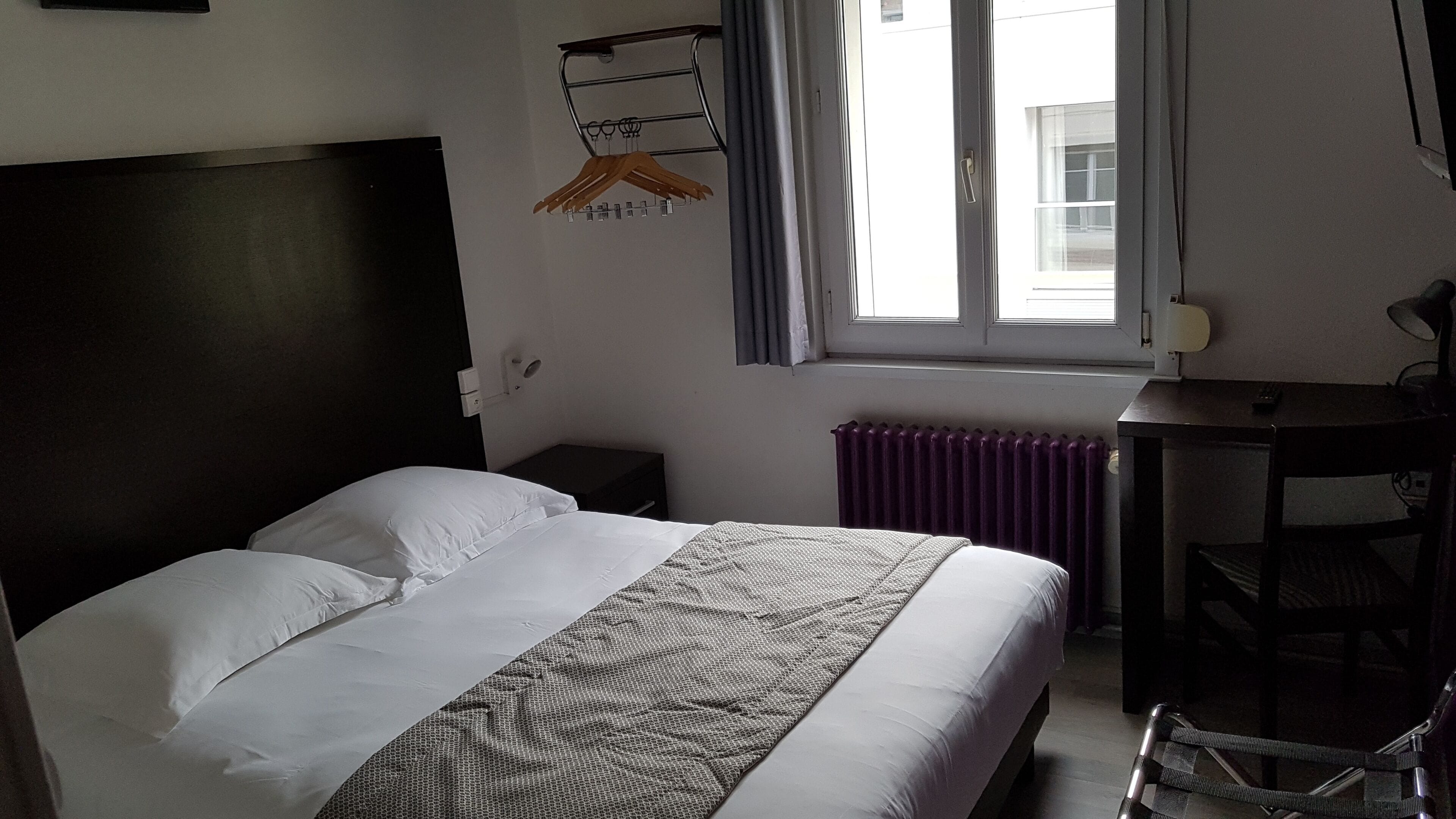 Una cama bien hecha con sábanas blancas y una manta estampada, una mesita de noche pequeña, una silla, un escritorio con una lámpara y una ventana con vistas a edificios.