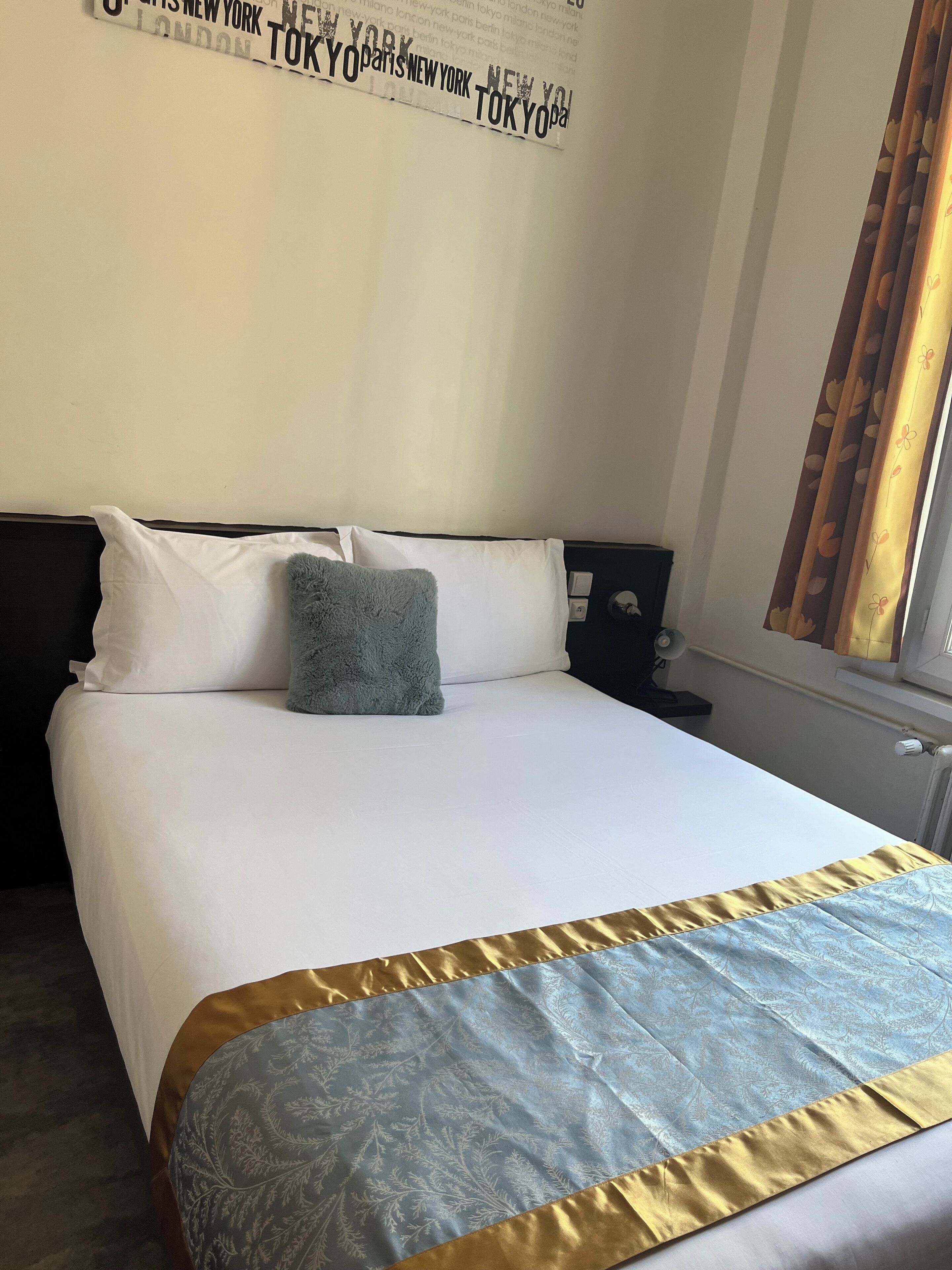Een net opgemaakt bed met een wit en gouden sprei, een zacht kussen en een muur met een naambord van een stad.