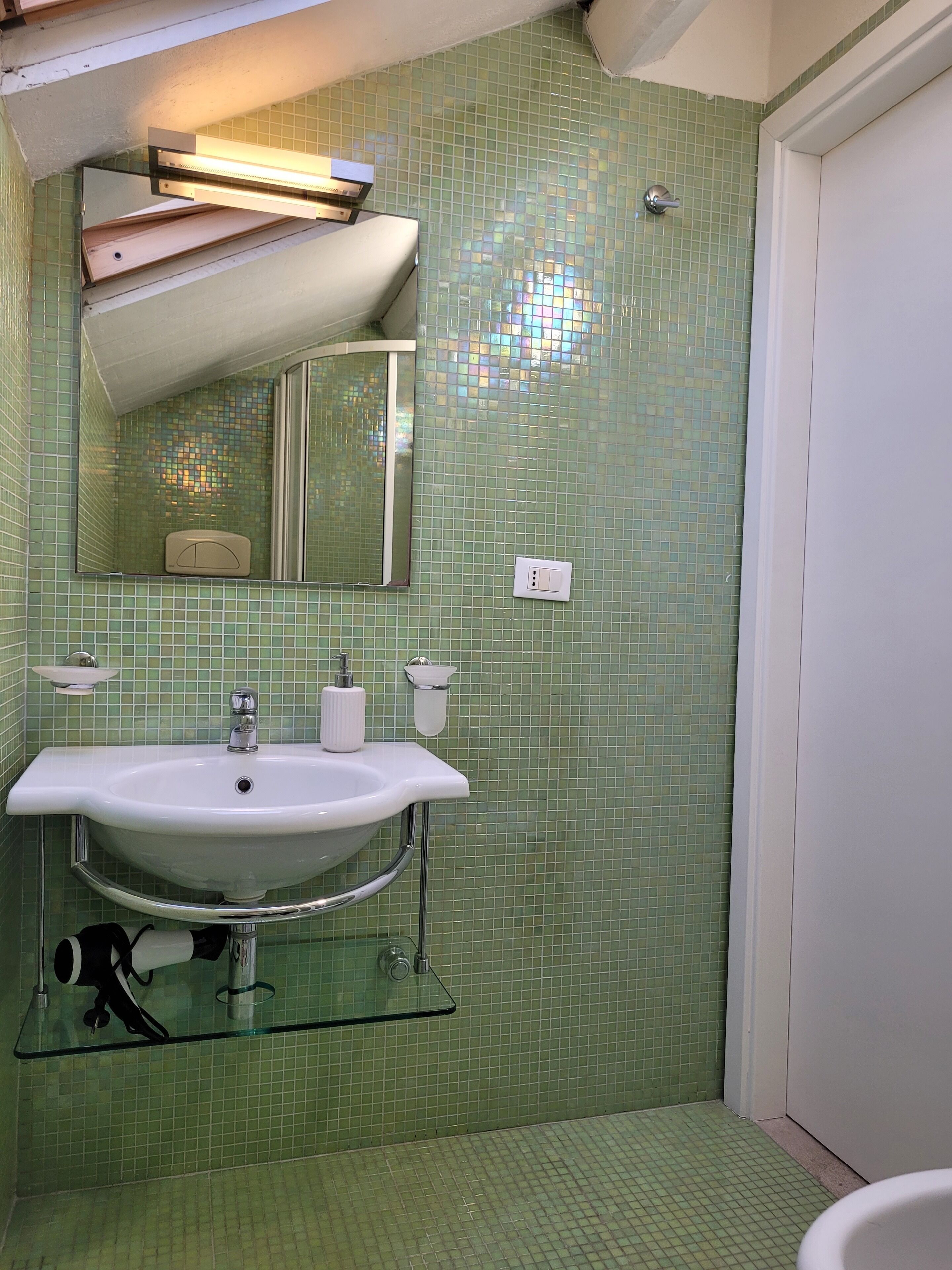 Une salle de bain avec un mur recouvert de carreaux verts, un lavabo blanc avec des robinets chromés, un miroir au-dessus du lavabo et une étagère en verre en dessous.