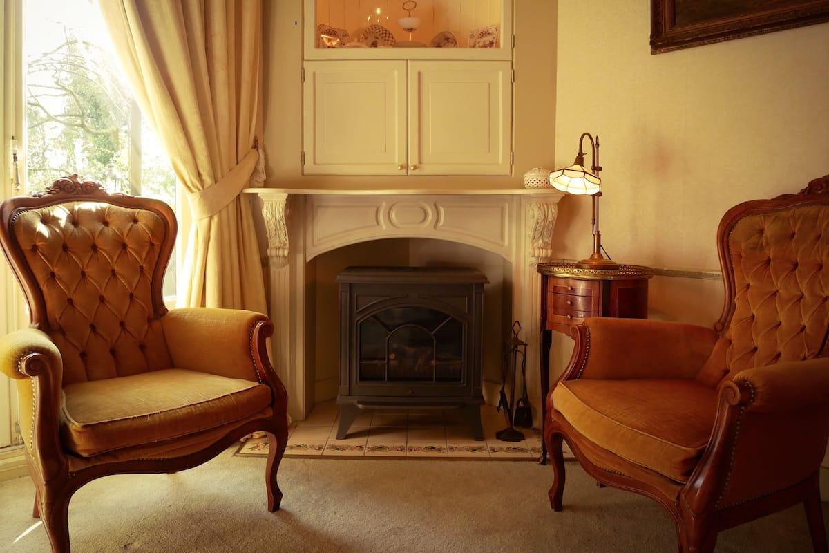 A room with a fireplace, two armchairs, a cabinet above the fireplace, and a lamp on a side table.