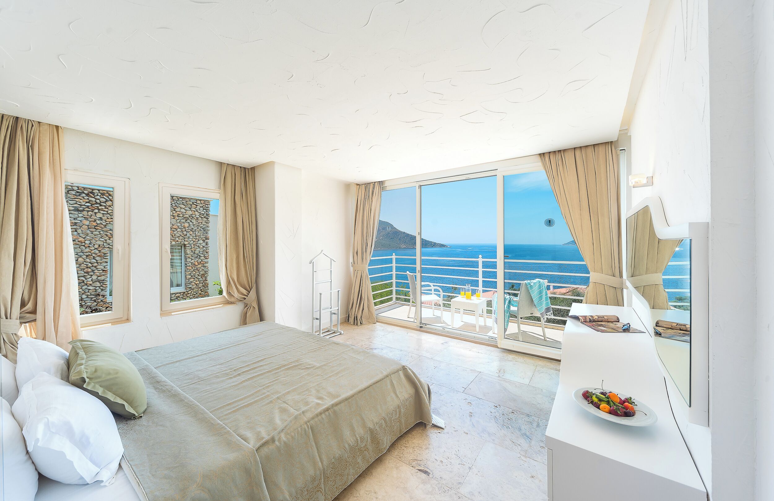 A bedroom with a bed, a balcony with a table and chairs, and a view of the sea.