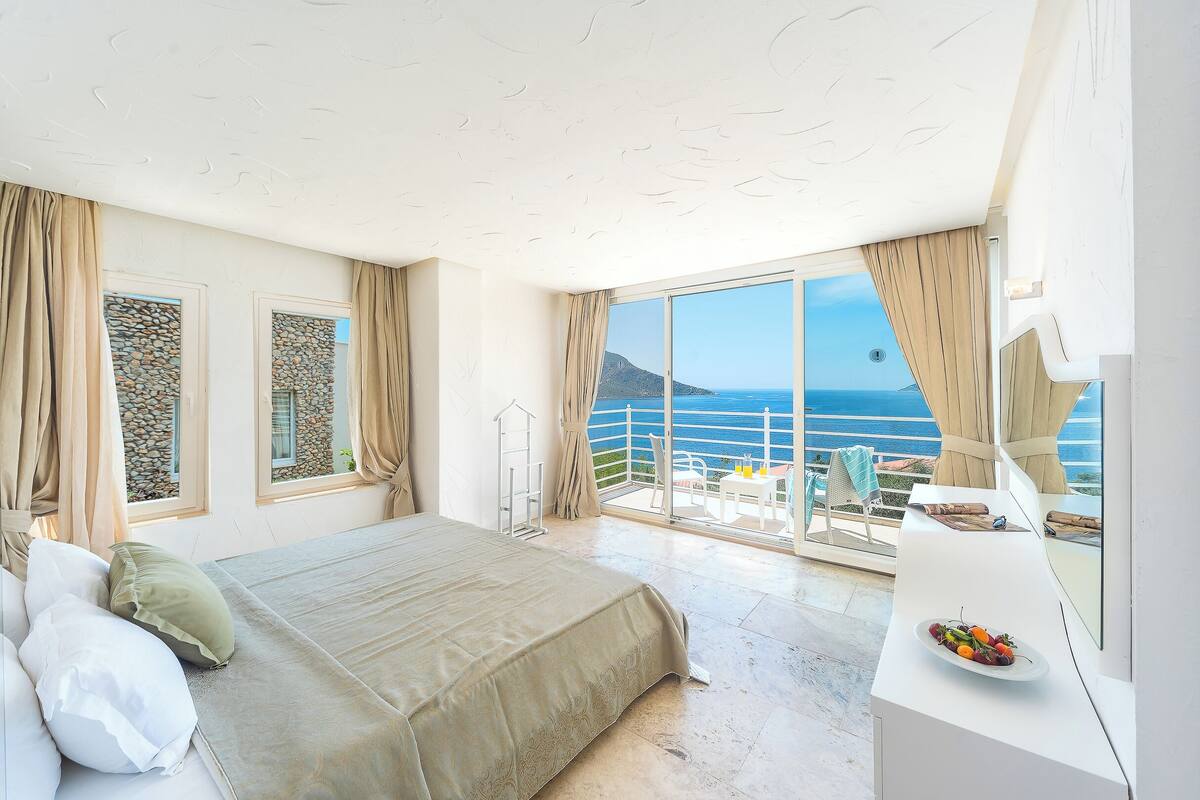 A bedroom with a bed, a balcony with a table and chairs, and a view of the sea.