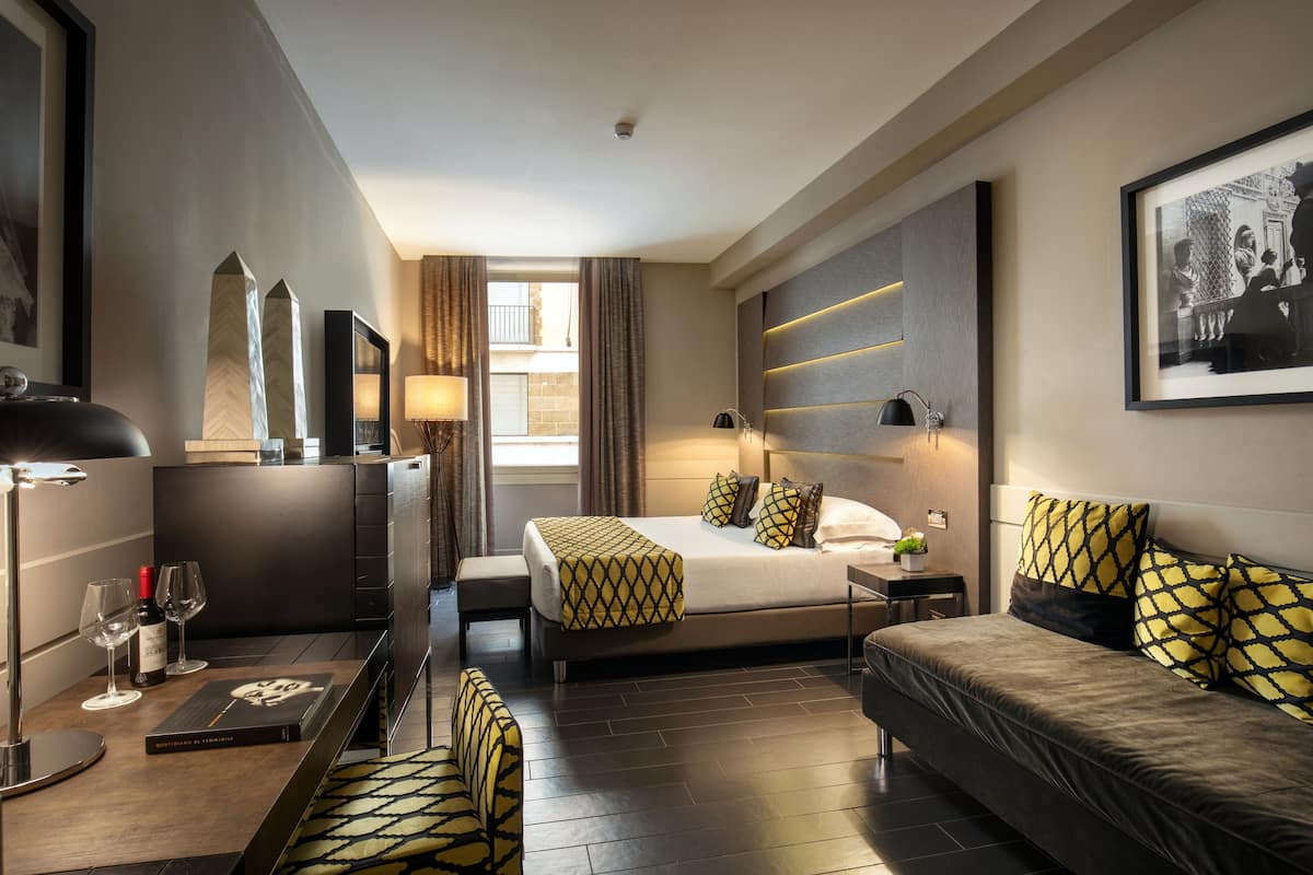 A modern hotel room with a large bed, a sofa, a desk with a lamp, and a view of the outside.