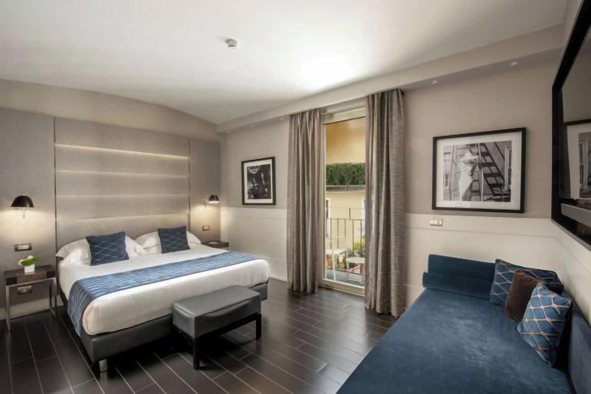 A modern hotel room with a large bed, a bench, a sofa, and a balcony with curtains.