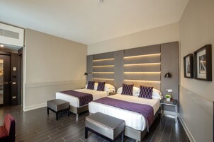A hotel room with two beds, a small table, a bench, and a nightstand with a lamp.