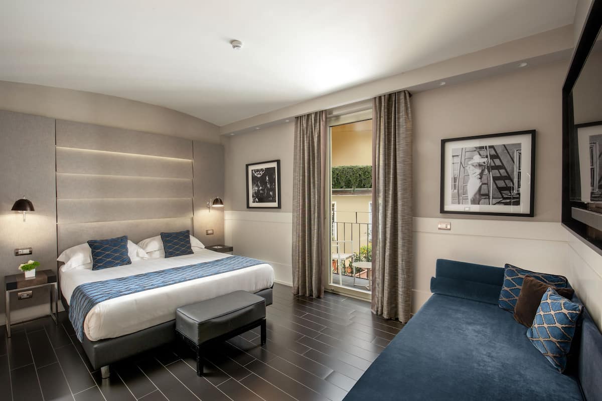 A modern hotel room with a large bed, a smaller sofa, a bench, and a balcony with curtains.