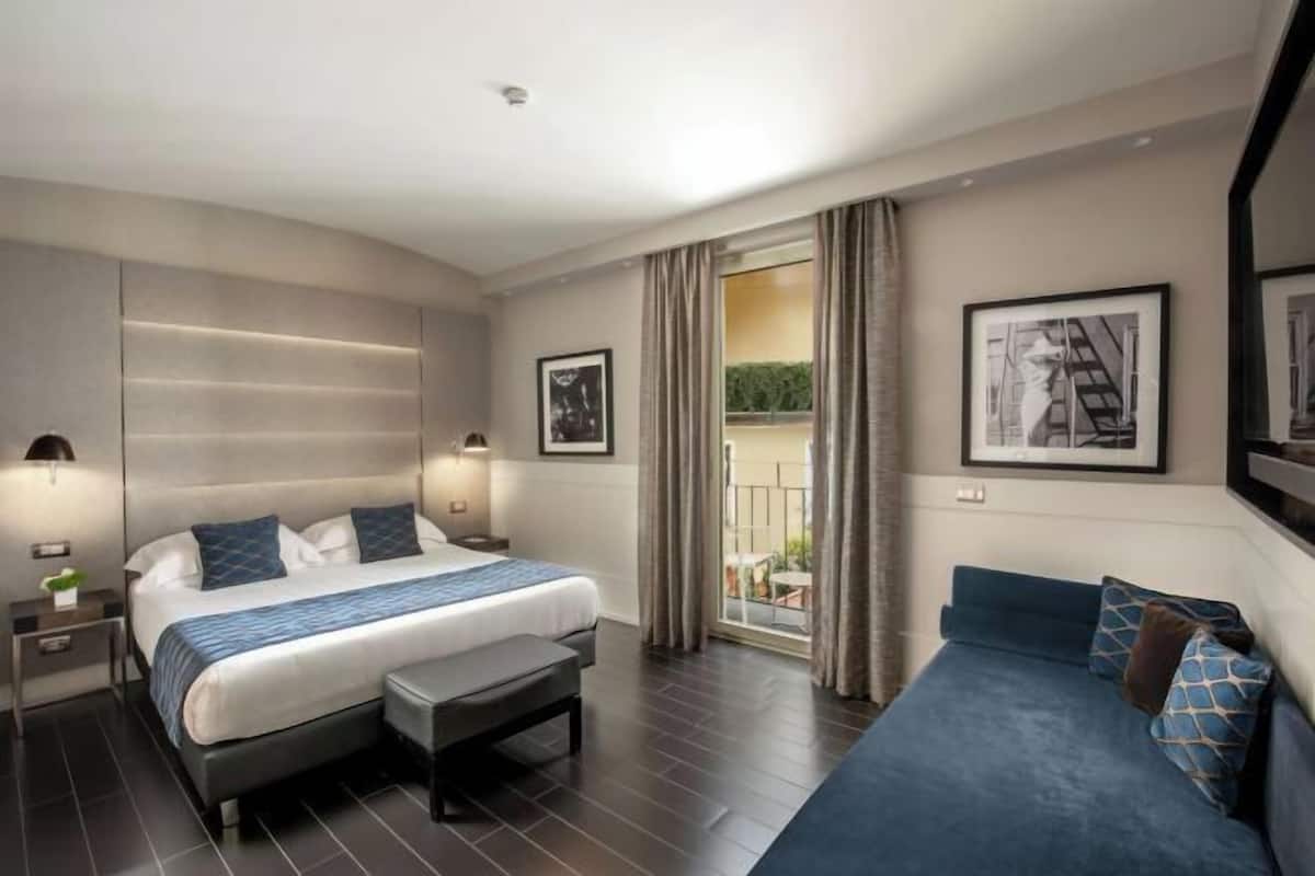 A modern hotel room with a large bed, a bench, a sofa, and a balcony with curtains.