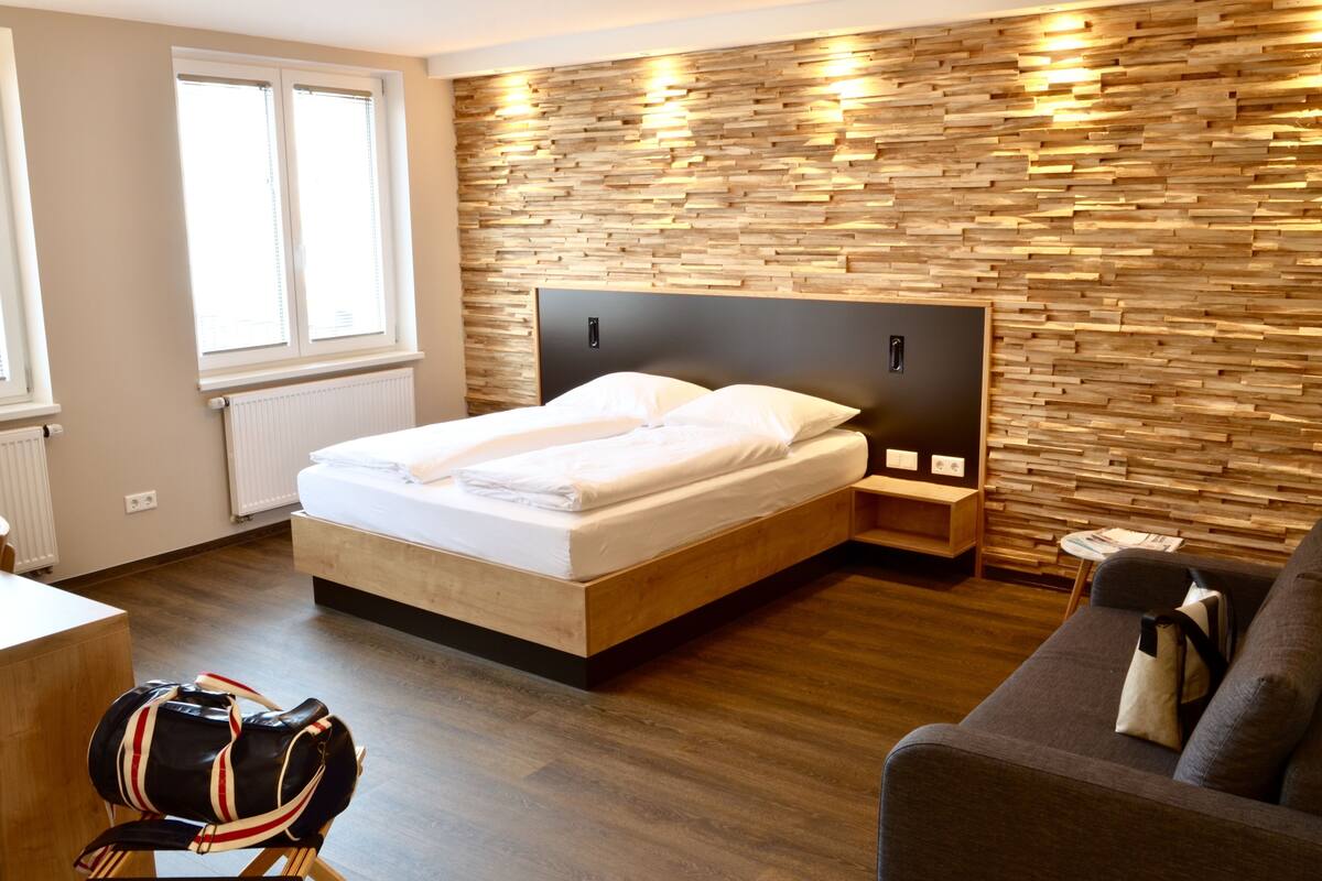 A modern bedroom with a wooden headboard, a bed with white linens, a wooden floor, a small bedside table with a lamp, and a window with blinds.