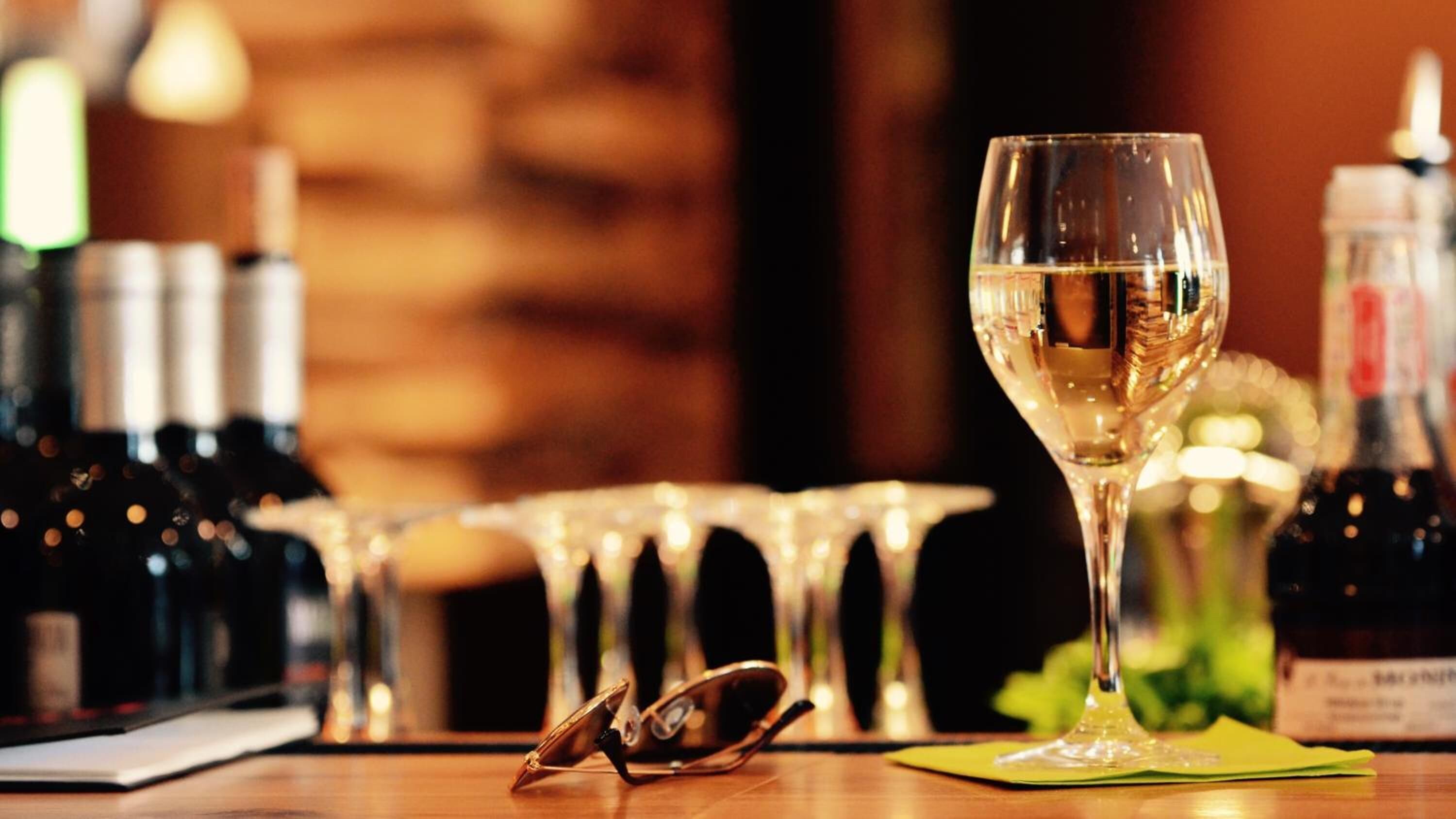 A bar setting with a glass of white wine, glasses lined up, and bottles of wine.