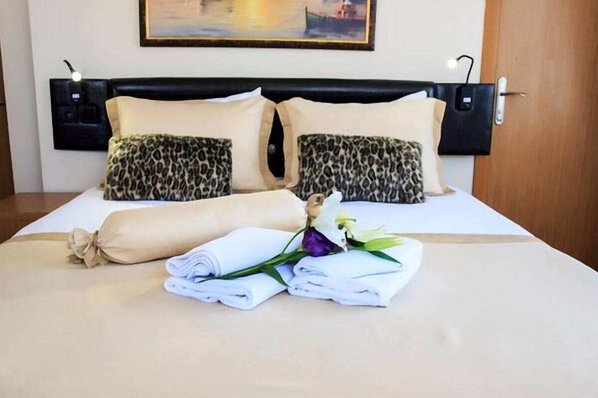 Una cama bien hecha con dos almohadones de estampado de leopardo, un almohadón color beige y una toalla blanca enrollada con una flor morada.