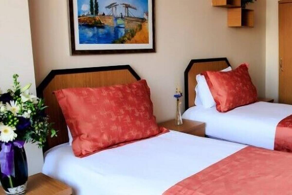 Habitación de hotel con dos camas, cada una con ropa de cama roja, cabeceros de madera, mesitas de noche, un jarrón de flores y un cuadro en la pared.