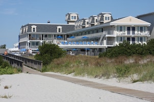 Un hotel frente a la playa con varios balcones, mobiliario exterior y un paseo marítimo que conduce a la playa.