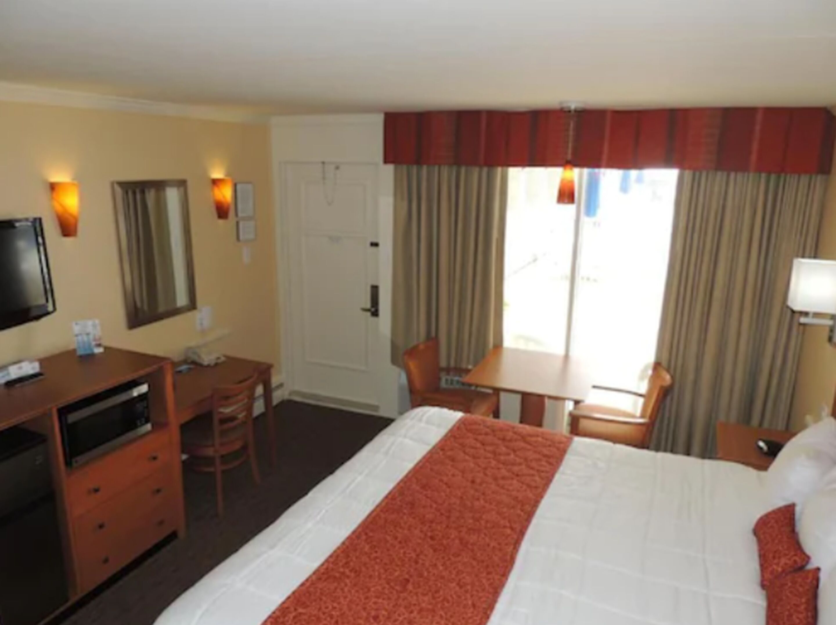 Una habitación de hotel con cama, escritorio, silla, televisor y ventana con cortinas.