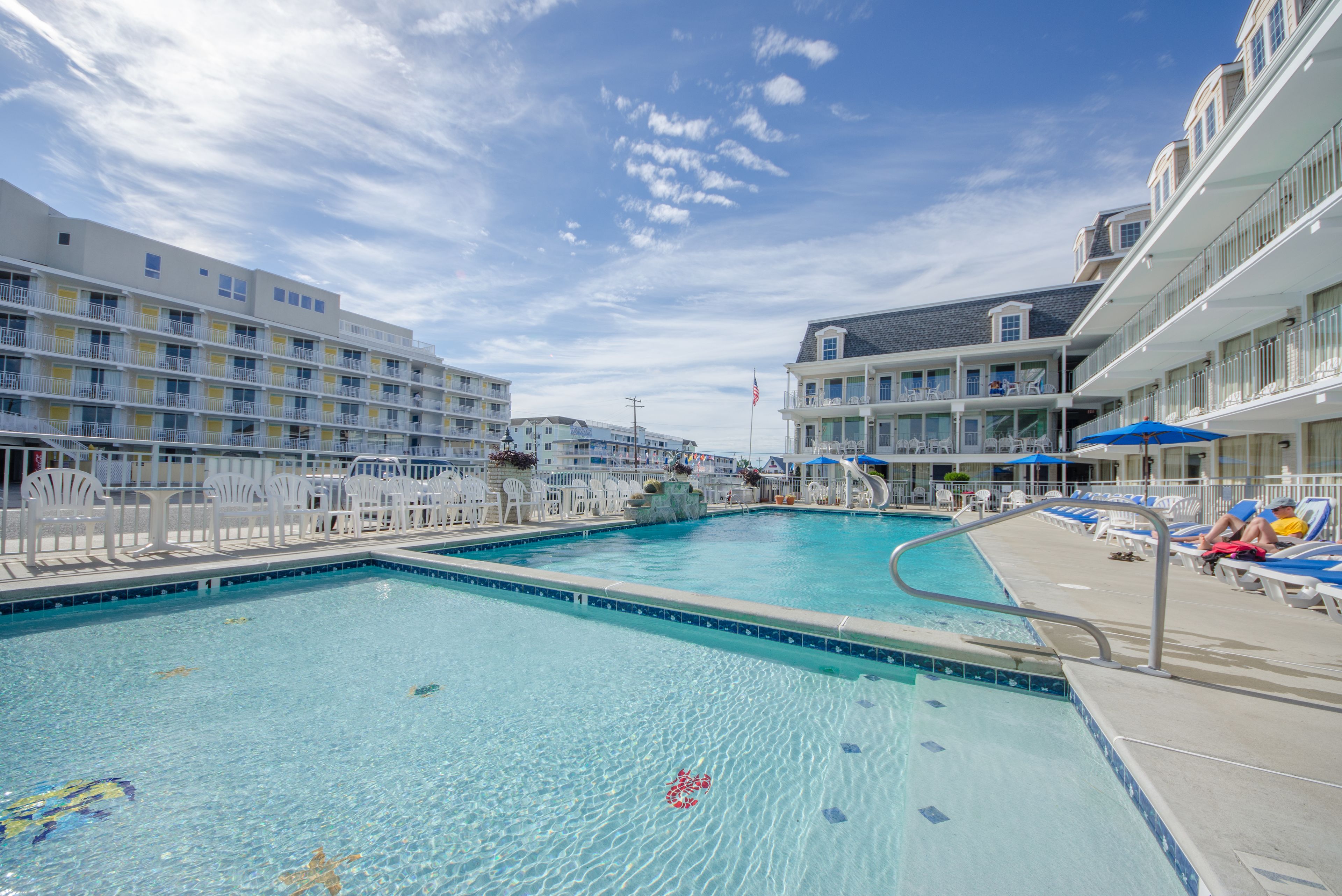 A swimming pool with a clear blue sky above, surrounded by white buildings with balconies and a flag.