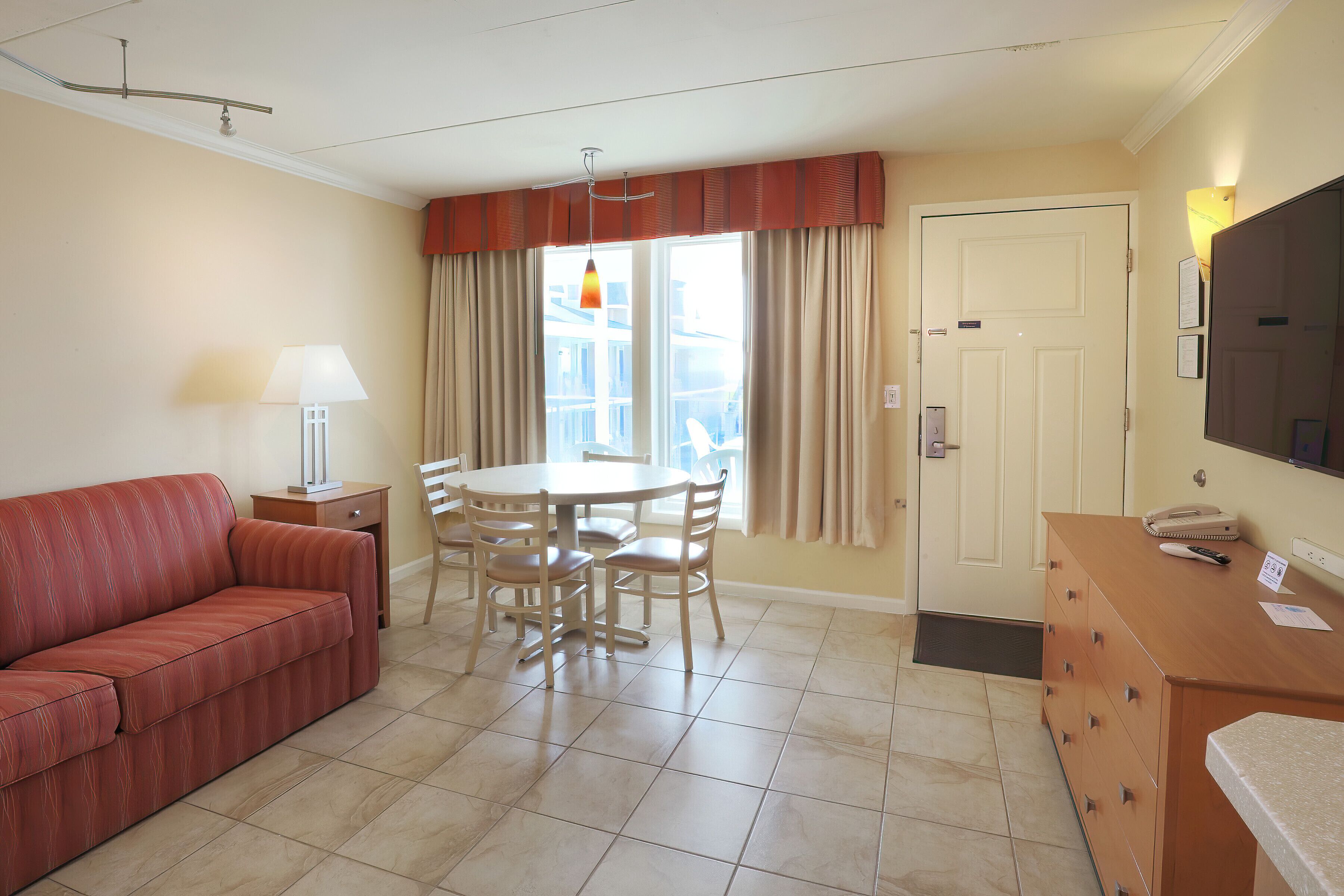 Ein Hotelzimmer mit einem roten Sofa, einem Esstisch mit Stühlen, einer Lampe, einem Fernseher und einer Tür mit Lichtschalter.