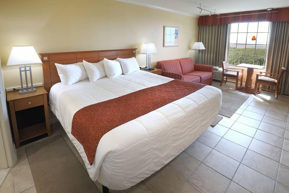 A hotel room with a large bed, two bedside tables with lamps, a chair, a small table, and a view of the outdoors through a window.