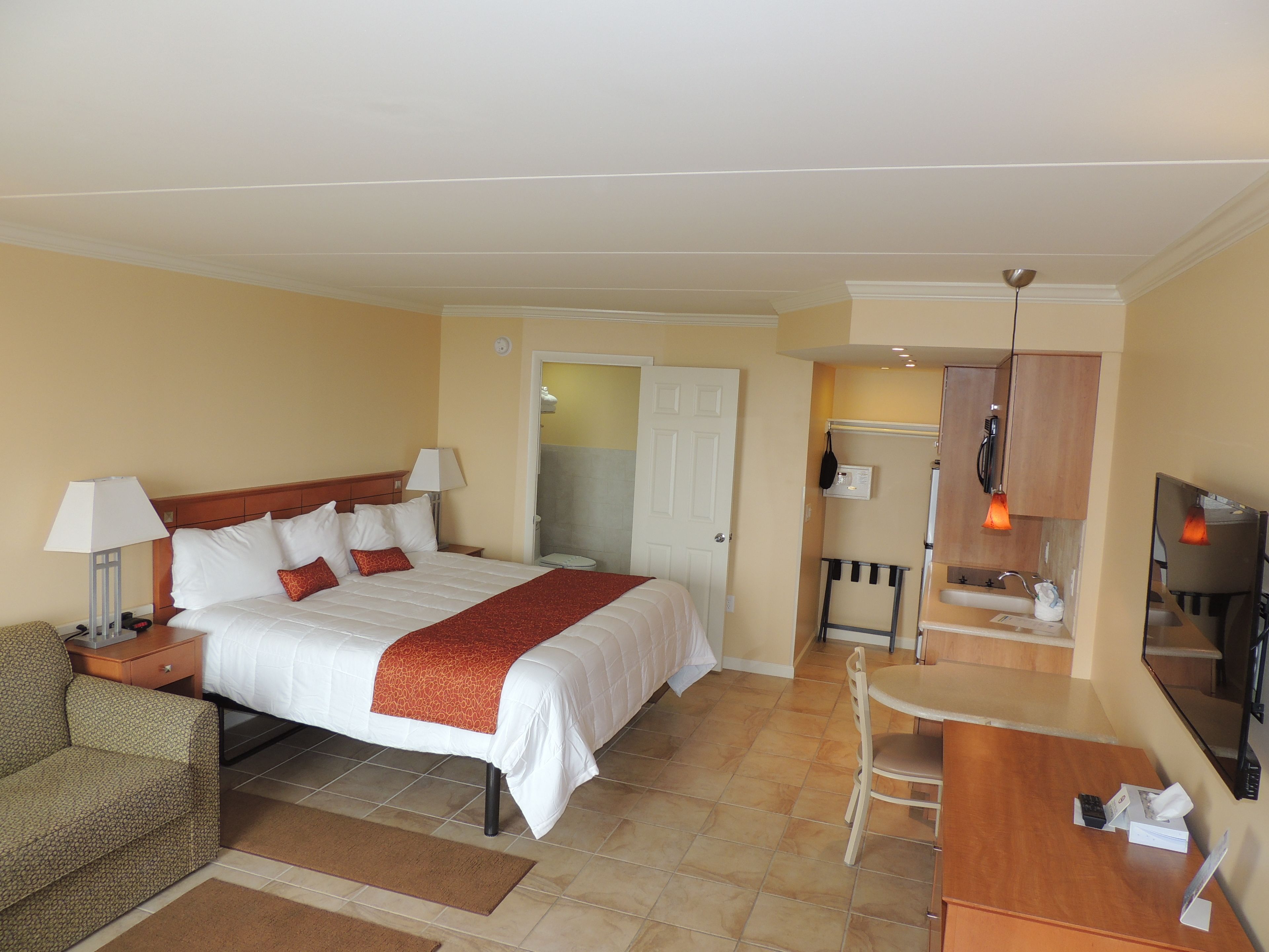A hotel room with a bed, bedside table, lamp, chair, desk, and a small kitchenette.