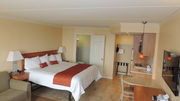 A hotel room with a bed, bedside table, lamp, chair, desk, and a small kitchenette.