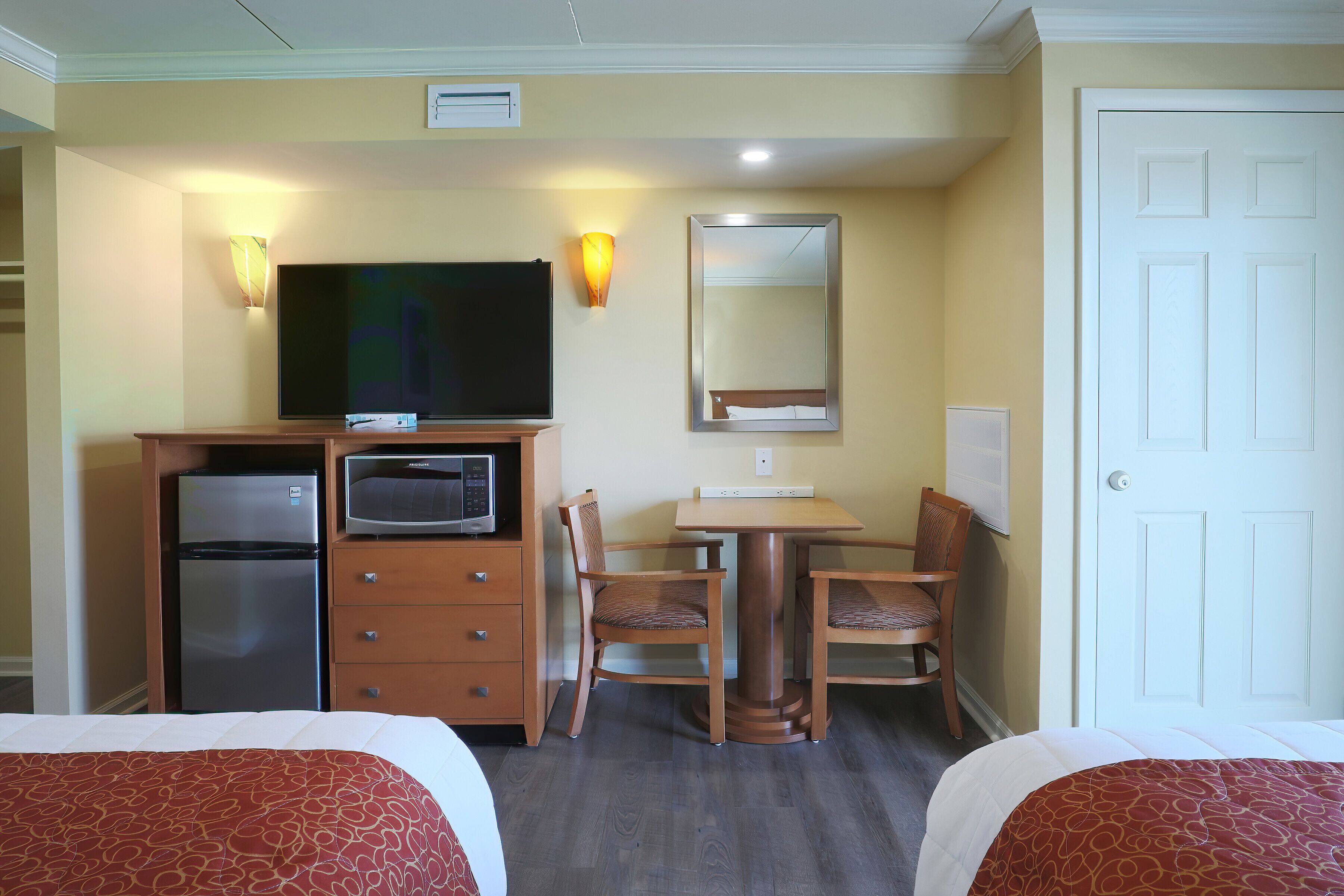 A hotel room with two beds, a desk with a television, a refrigerator, and a dining table with chairs.