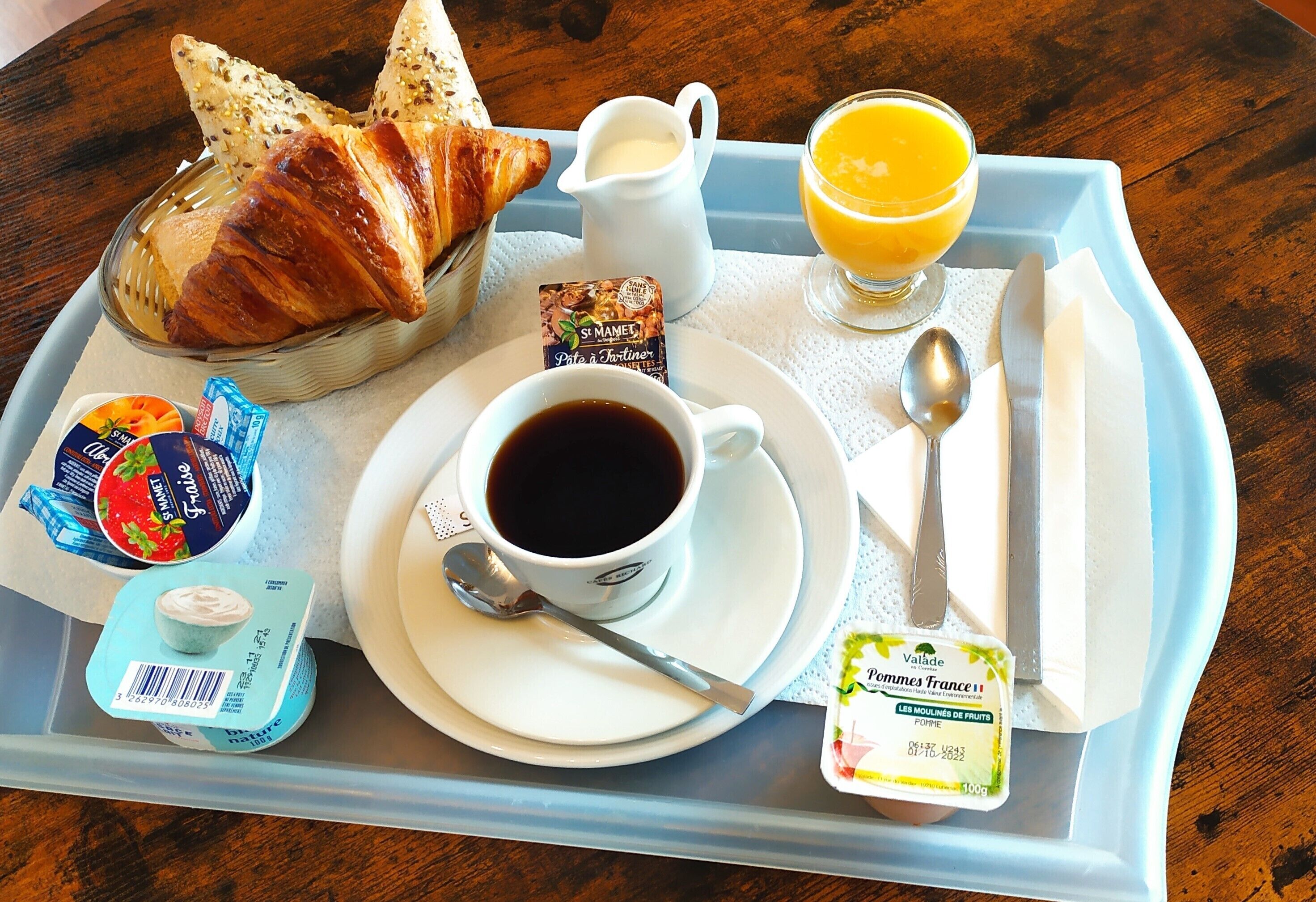 A breakfast tray with croissants, a jug of milk, a glass of orange juice, a cup of coffee, yogurt, and a packet of fruit mueslins.